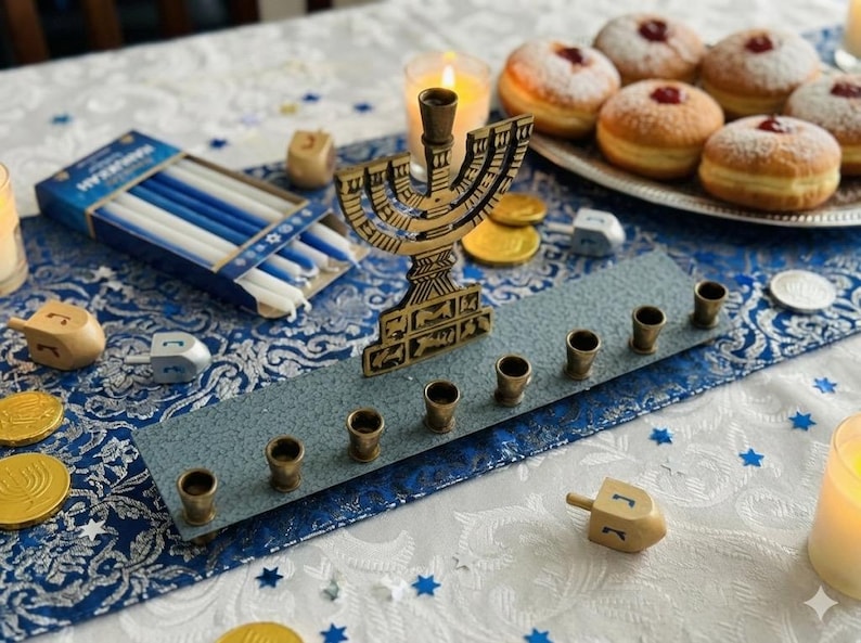 May include: A Hanukkah display with a brass menorah, candles, dreidels, and sufganiyot. The menorah sits on a blue and grey decorative tray. Gold coins and blue and white candles are also present. The table is decorated with blue stars.