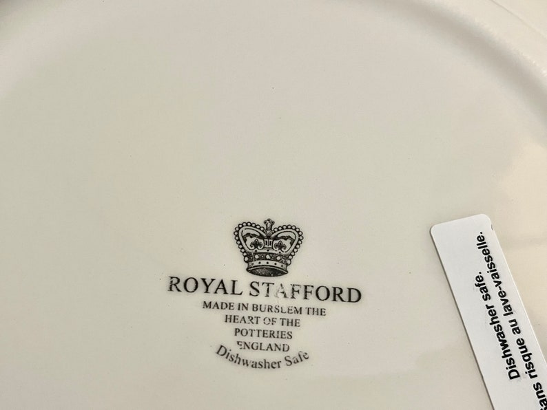 Może przedstawiać: Biały ceramiczny talerz z czarno-białym logo Royal Stafford. Logo zawiera koronę i tekst "Royal Stafford" oraz "Made in Burslem the heart of the potteries England Dishwasher Safe".