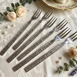 May include: Six vintage silver forks with ornate floral designs on the handles. The forks are arranged on a white tablecloth with floral accents, pearls, and a teacup in the background. A classic dining set.