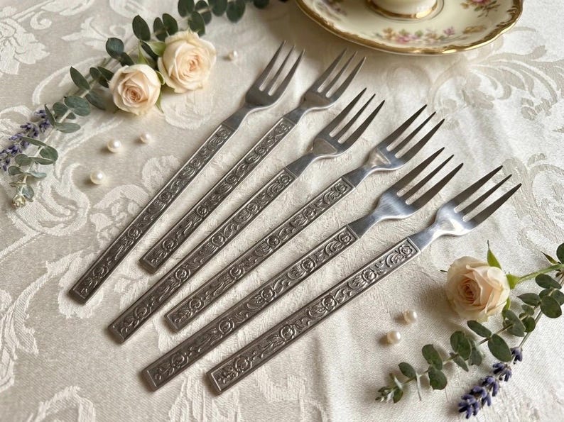 May include: Six vintage silver forks with ornate floral designs on the handles. The forks are arranged on a white tablecloth with floral accents, pearls, and a teacup in the background. A classic dining set.