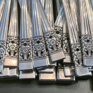 May include: A collection of silver-toned metal flatware handles arranged in a cluster. Each handle features a vertical ribbed pattern along the stem and a detailed embossed floral motif at the base, characterized by intricate petals and leaves.