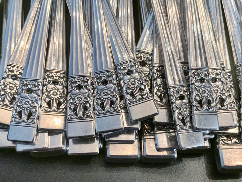 May include: A collection of silver-toned metal flatware handles arranged in a cluster. Each handle features a vertical ribbed pattern along the stem and a detailed embossed floral motif at the base, characterized by intricate petals and leaves.