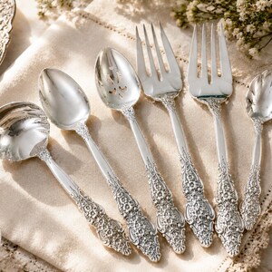 May include: A set of six silver-toned metal utensils featuring intricate floral embossed handles. The collection includes a round soup spoon, a standard spoon, a pierced spoon, two forks of varying sizes, and a small scalloped-edge spoon.