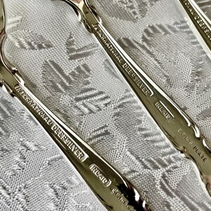 May include: Close-up of several silver spoons with ornate handles. The spoons are marked "International DeepSilver Inlaid E.P.N.S. Plate". The spoons are arranged on a white patterned tablecloth, reflecting the light.