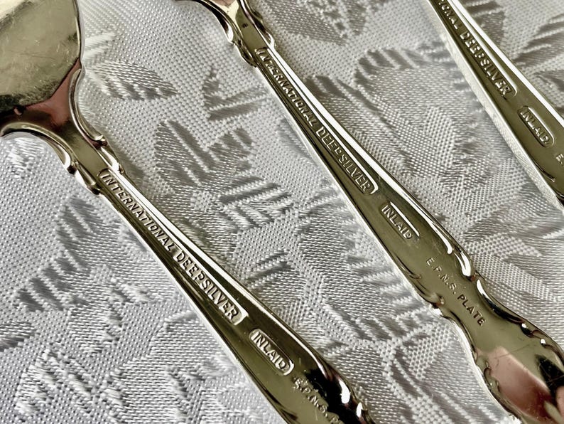 May include: Close-up of several silver spoons with ornate handles. The spoons are marked "International DeepSilver Inlaid E.P.N.S. Plate". The spoons are arranged on a white patterned tablecloth, reflecting the light.