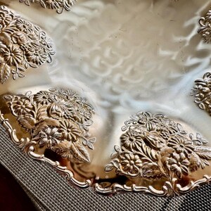 May include: Silver-toned decorative serving tray with a scalloped edge. The tray features embossed floral designs around the rim and in the corners. The reflective surface of the tray creates a bright, elegant appearance.
