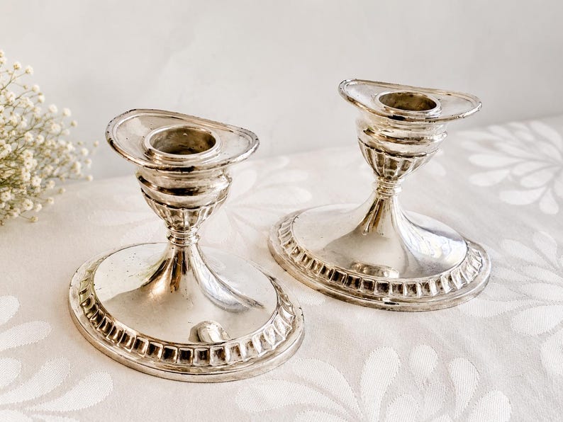 Pu&ograve; includere: Due portacandele in metallo color argento con basi ovali e stelo scanalato. Ogni pezzo presenta un bordo largo e piatto attorno all'alloggiamento centrale per la candela e un motivo decorativo lungo il bordo della base ovale, riflettendo la luce.