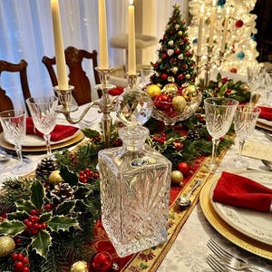 May include: A festive Christmas table setting with a crystal decanter, lit candles, and a small Christmas tree. The table is decorated with holly, ornaments, and gold accents. Wine glasses and plates are set for a holiday meal.