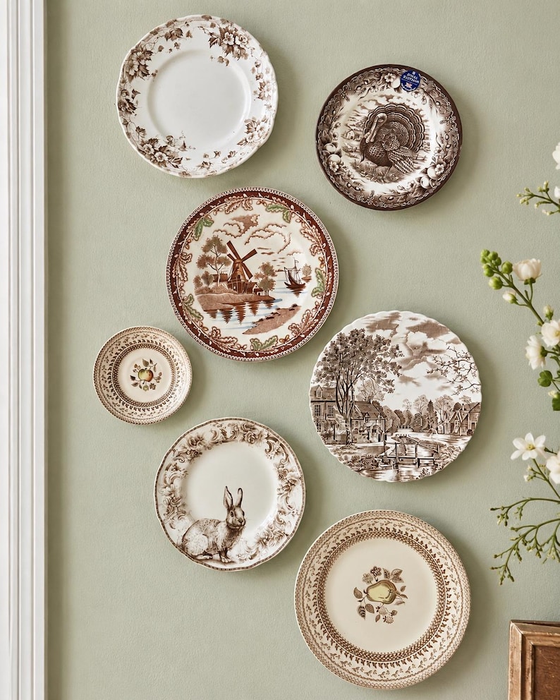May include: A collection of six round ceramic plates displayed on a wall. The plates feature brown and white transferware patterns including a turkey, a windmill landscape, a rabbit, a pear, an apple, and a detailed village scene with trees.