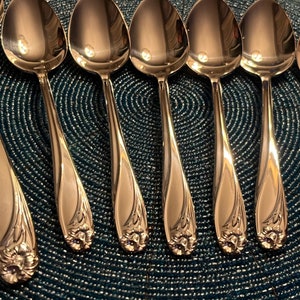 May include: A set of polished silver spoons arranged on a textured blue surface. Each spoon features a decorative floral design at the base of the handle. The spoons are reflective, showing the surrounding environment.
