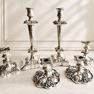May include: A collection of six silver-toned metal candle holders featuring ornate embossed grape and vine motifs on the bases and stems. The set includes two tall, slender candlesticks and four shorter, wider candle stands with decorative edges.