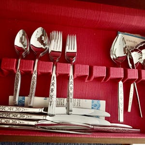 May include: A set of stainless steel flatware arranged in a red storage case. The collection includes spoons, forks, knives, and serving utensils, each featuring a decorative floral pattern on the handle. Some pieces are upright in slots.