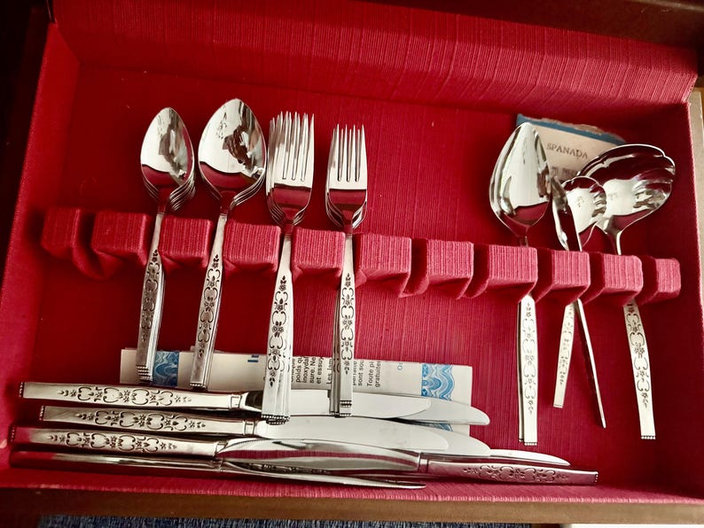 May include: A set of stainless steel flatware arranged in a red storage case. The collection includes spoons, forks, knives, and serving utensils, each featuring a decorative floral pattern on the handle. Some pieces are upright in slots.