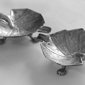 K&ouml;nnte beinhalten: Zwei silberfarbene, dekorative Aschenbecher. Jeder Aschenbecher ist wie ein F&auml;cher geformt und mit aufwendigen ge&auml;tzten Designs versehen. Sie haben kleine, krallenartige F&uuml;&szlig;e und eine gebogene Ablage f&uuml;r eine Zigarette oder Zigarre. Die Aschenbecher werden auf einer hellen Oberfl&auml;che pr&auml;sentiert.