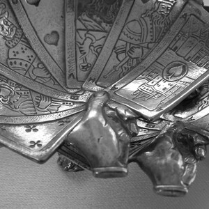 K&ouml;nnte beinhalten: Ein silberfarbenes Dekorationsobjekt, das eine Hand zeigt, die ein aufgef&auml;chertes Kartenspiel h&auml;lt. Die Karten zeigen detaillierte Illustrationen von K&ouml;nigen, K&ouml;niginnen und Kartensymbolen. Wahrscheinlich ein Dekorationsartikel oder Kartenhalter.