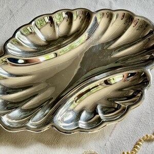Pode incluir: Um prato de servir em metal prateado polido com a forma de uma grande concha de vieira. O prato apresenta uma superf&iacute;cie lisa e refletora com sulcos curvos profundos que irradiam do centro para as bordas recortadas, criando uma forma de concha.