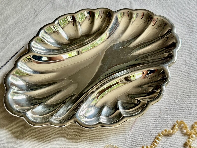 Pode incluir: Um prato de servir em metal prateado polido com a forma de uma grande concha de vieira. O prato apresenta uma superf&iacute;cie lisa e refletora com sulcos curvos profundos que irradiam do centro para as bordas recortadas, criando uma forma de concha.
