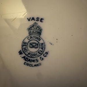 K&ouml;nnte beinhalten: Nahaufnahme einer wei&szlig;en Porzellanvase mit blauem Emblem. Das Emblem zeigt eine Krone &uuml;ber einem kreisf&ouml;rmigen Design mit den Worten "SEMI-PORCELAIN" und "W. ADAMS & CO. ENGLAND". Das Wort "VASE" ist &uuml;ber der Krone aufgedruckt.