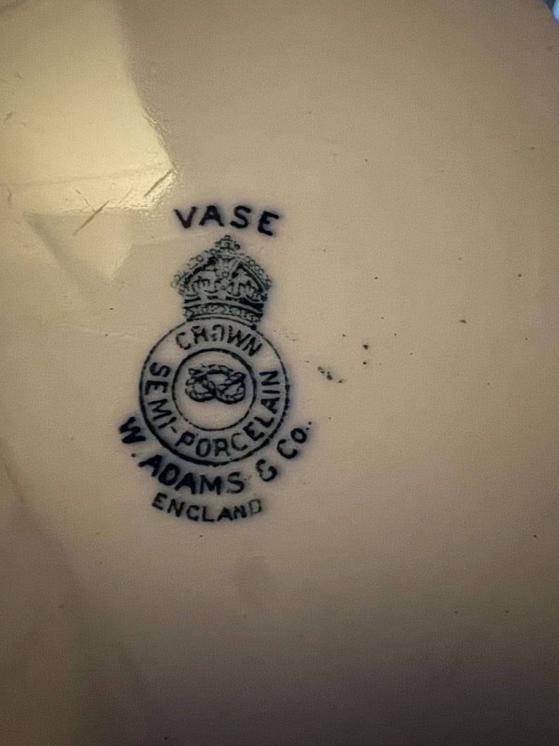 K&ouml;nnte beinhalten: Nahaufnahme einer wei&szlig;en Porzellanvase mit blauem Emblem. Das Emblem zeigt eine Krone &uuml;ber einem kreisf&ouml;rmigen Design mit den Worten "SEMI-PORCELAIN" und "W. ADAMS & CO. ENGLAND". Das Wort "VASE" ist &uuml;ber der Krone aufgedruckt.