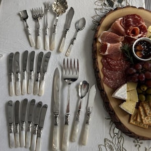 May include: A collection of vintage silverware with mother-of-pearl handles, including forks, knives, and spoons, displayed on a white tablecloth. A wooden serving board presents charcuterie, cheese, grapes, and crackers.