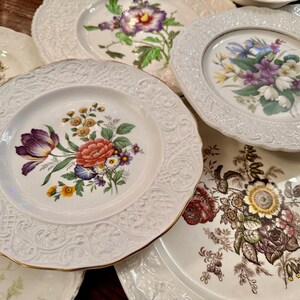 May include: A collection of white ceramic plates featuring intricate embossed rims and central floral illustrations. The designs include various colorful bouquets with red, purple, yellow, and green flowers, finished with delicate gold-toned edges.