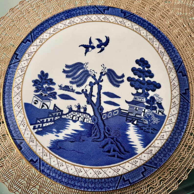 May include: A blue and white porcelain plate with a willow tree design. The plate has a gold rim and a gold pattern around the edge.