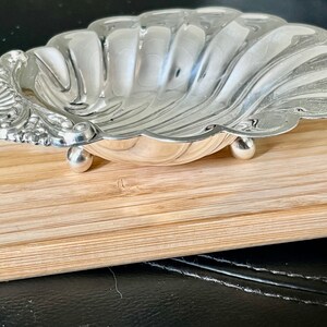 May include: A silver shell-shaped dish with a decorative handle and three feet. The dish is sitting on a wooden surface.