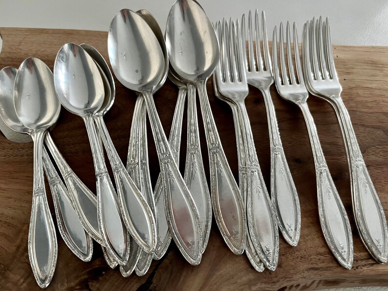 May include: A set of silver-plated spoons and forks with ornate designs. The silverware is arranged on a wooden surface.