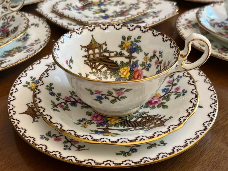 K&ouml;nnte beinhalten: Ein wei&szlig;es Tee-Set mit Blumenmuster und goldener Verzierung. Die Teetasse zeigt ein zartes Blumenmuster mit einem Pagoden- und Br&uuml;ckenmotiv. Die Untertasse hat ein passendes Blumenmuster mit einem goldenen Rand.