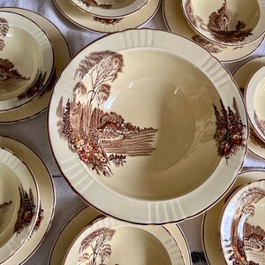 May include: A set of vintage cream-coloured ceramic bowls with brown floral designs. The bowls feature a cottage scene with trees, a house, and a path.