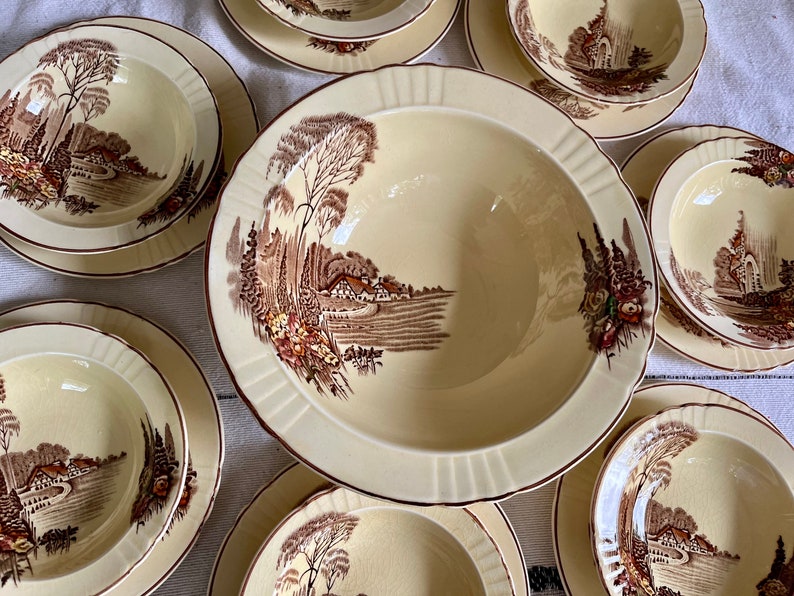 May include: A set of vintage cream-coloured ceramic bowls with brown floral designs. The bowls feature a cottage scene with trees, a house, and a path.