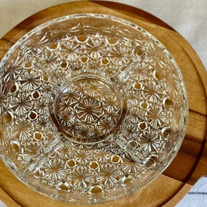 May include: A clear glass serving dish with a starburst pattern sits atop a round wooden serving board with a handle. The glass dish is divided into sections, perfect for serving snacks or appetizers. The wooden board has a natural wood grain.