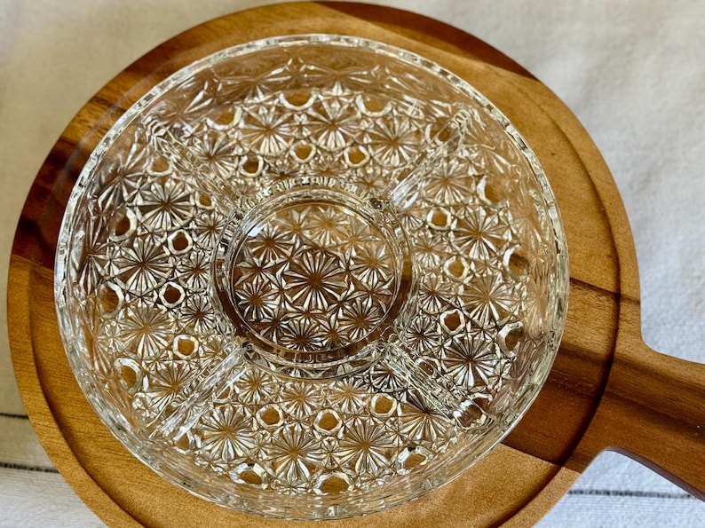 May include: A clear glass serving dish with a starburst pattern sits atop a round wooden serving board with a handle. The glass dish is divided into sections, perfect for serving snacks or appetizers. The wooden board has a natural wood grain.