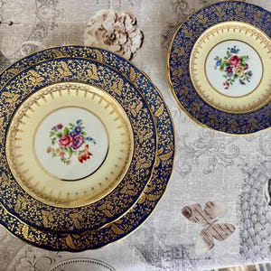 May include: A set of fine china plates with a floral design. The plates have a cream-colored center with a bouquet of flowers and a navy blue border with gold accents. The set includes plates of different sizes.