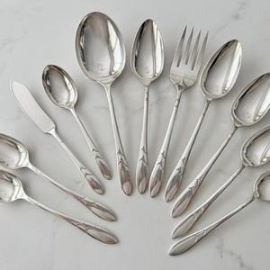May include: A collection of ten silver-toned metal utensils arranged in a fan shape. The set includes eight spoons of varying sizes, one fork with four tines, and one flat-bladed knife. Each handle features a delicate embossed floral motif.