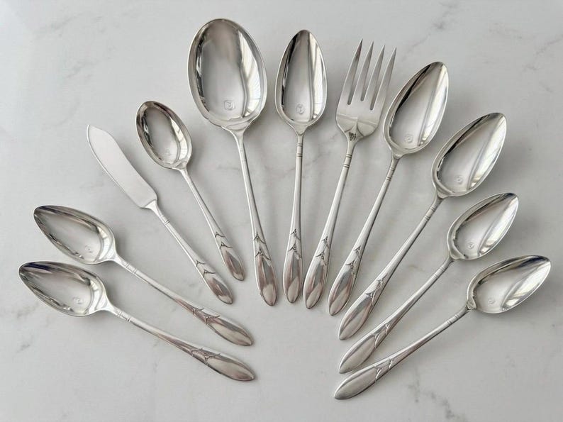 May include: A collection of ten silver-toned metal utensils arranged in a fan shape. The set includes eight spoons of varying sizes, one fork with four tines, and one flat-bladed knife. Each handle features a delicate embossed floral motif.
