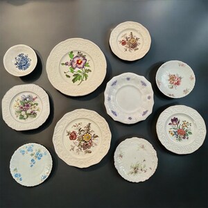 May include: A collection of nine ceramic plates and bowls of varying sizes, featuring white backgrounds with embossed rims and colorful floral patterns, including depictions of purple, yellow, red, and blue flowers, arranged on a dark surface.