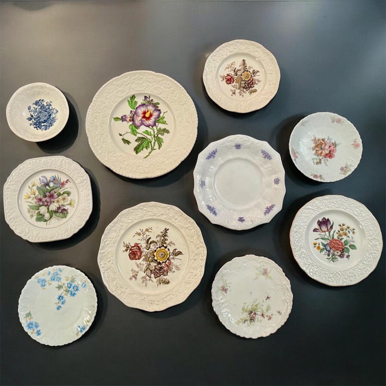May include: A collection of nine ceramic plates and bowls of varying sizes, featuring white backgrounds with embossed rims and colorful floral patterns, including depictions of purple, yellow, red, and blue flowers, arranged on a dark surface.
