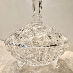 May include: A clear, cut-glass candy dish featuring a multifaceted starburst pattern across the bowl and lid. The vessel is supported by three small, integrated feet and is topped with a pointed, faceted finial handle on the removable lid.