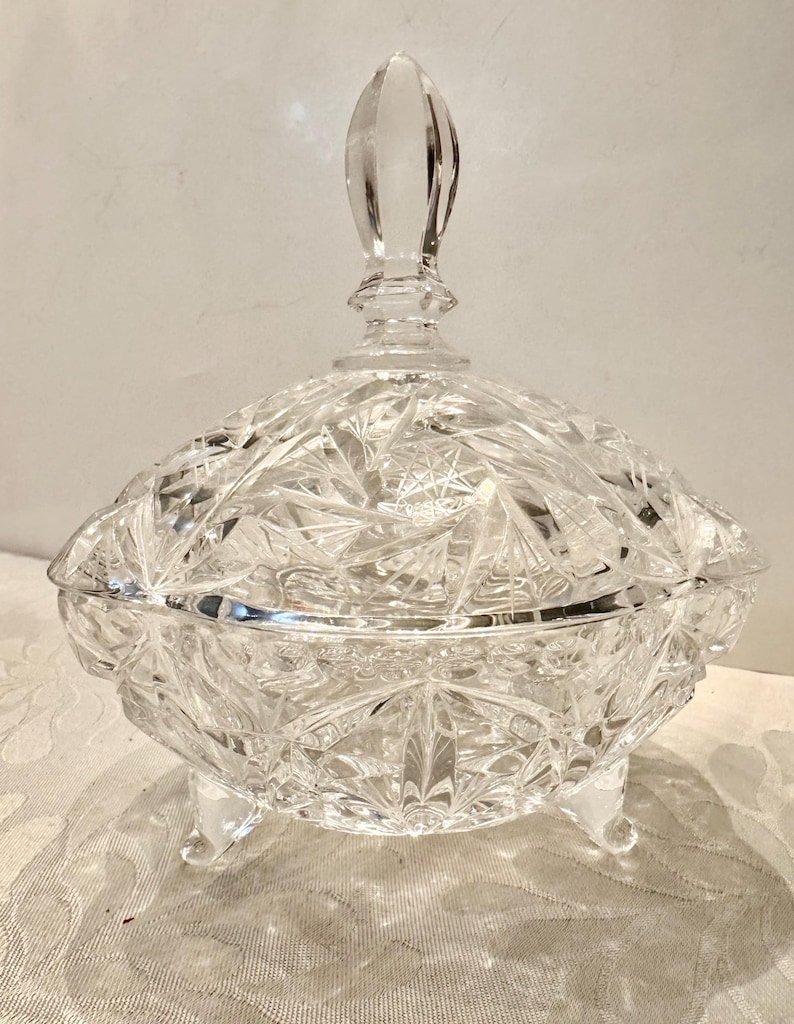 May include: A clear, cut-glass candy dish featuring a multifaceted starburst pattern across the bowl and lid. The vessel is supported by three small, integrated feet and is topped with a pointed, faceted finial handle on the removable lid.