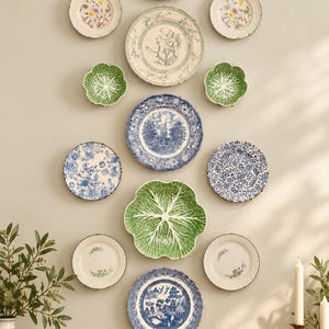 May include: A collection of twelve decorative ceramic plates and bowls arranged on a wall. The set includes blue and white transferware, floral patterned plates, and green cabbage-shaped bowls, creating a varied display of dinnerware.