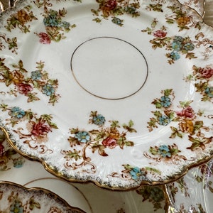 K&ouml;nnte beinhalten: Set aus drei wei&szlig;en Porzellantellern mit goldfarbenem Rand und Blumenmuster. Die Teller haben einen gewellten Rand und sind mit zarten rosa, blauen und gelben Blumen verziert.