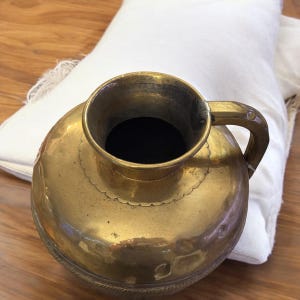 K&ouml;nnte beinhalten: Eine gro&szlig;e, antike Messingkanne mit einer breiten &Ouml;ffnung und einem einzigen Henkel. Die Kanne hat einen dekorativen, gewellten Rand und komplizierte Details.