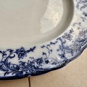 Puede incluir: Plato ovalado de cer&aacute;mica blanca con un dise&ntilde;o floral azul alrededor del borde. El patr&oacute;n presenta flores y follaje detallados. El plato tiene un borde ligeramente festoneado y un borde azul oscuro.