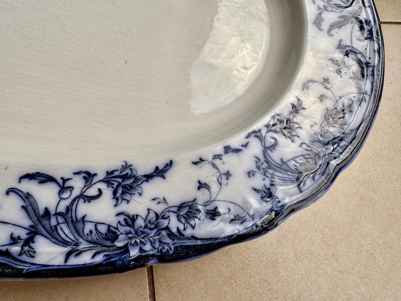 Puede incluir: Plato ovalado de cer&aacute;mica blanca con un dise&ntilde;o floral azul alrededor del borde. El patr&oacute;n presenta flores y follaje detallados. El plato tiene un borde ligeramente festoneado y un borde azul oscuro.