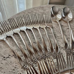 May include: A set of silver flatware, including forks and spoons, with ornate handles. The flatware is arranged on a white lace tablecloth.