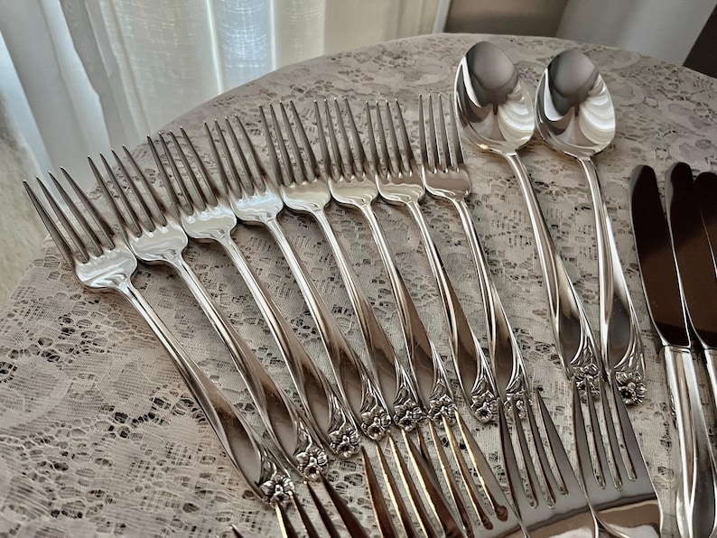 May include: A set of silver flatware, including forks and spoons, with ornate handles. The flatware is arranged on a white lace tablecloth.