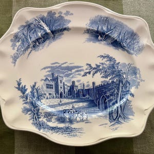 Op de afbeelding: Een ovale witte keramische serveerschaal met een geschulpte rand, voorzien van blauwe transferware-illustraties van een groot stenen landhuis, een paardenkoets en omliggende landschapssc&egrave;nes met bomen en architectonische details.