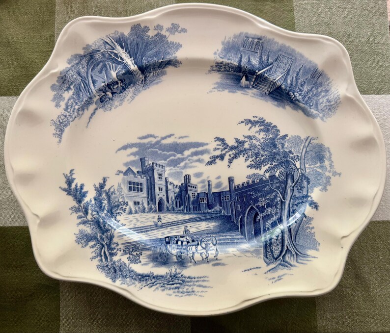 Op de afbeelding: Een ovale witte keramische serveerschaal met een geschulpte rand, voorzien van blauwe transferware-illustraties van een groot stenen landhuis, een paardenkoets en omliggende landschapssc&egrave;nes met bomen en architectonische details.