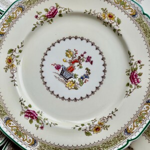Puede incluir: Un plato de cer&aacute;mica blanco con un borde verde y dorado. El plato presenta un dise&ntilde;o floral con flores rosas, amarillas y verdes. El centro del plato tiene un dise&ntilde;o floral m&aacute;s peque&ntilde;o con una maceta marr&oacute;n y una planta verde.